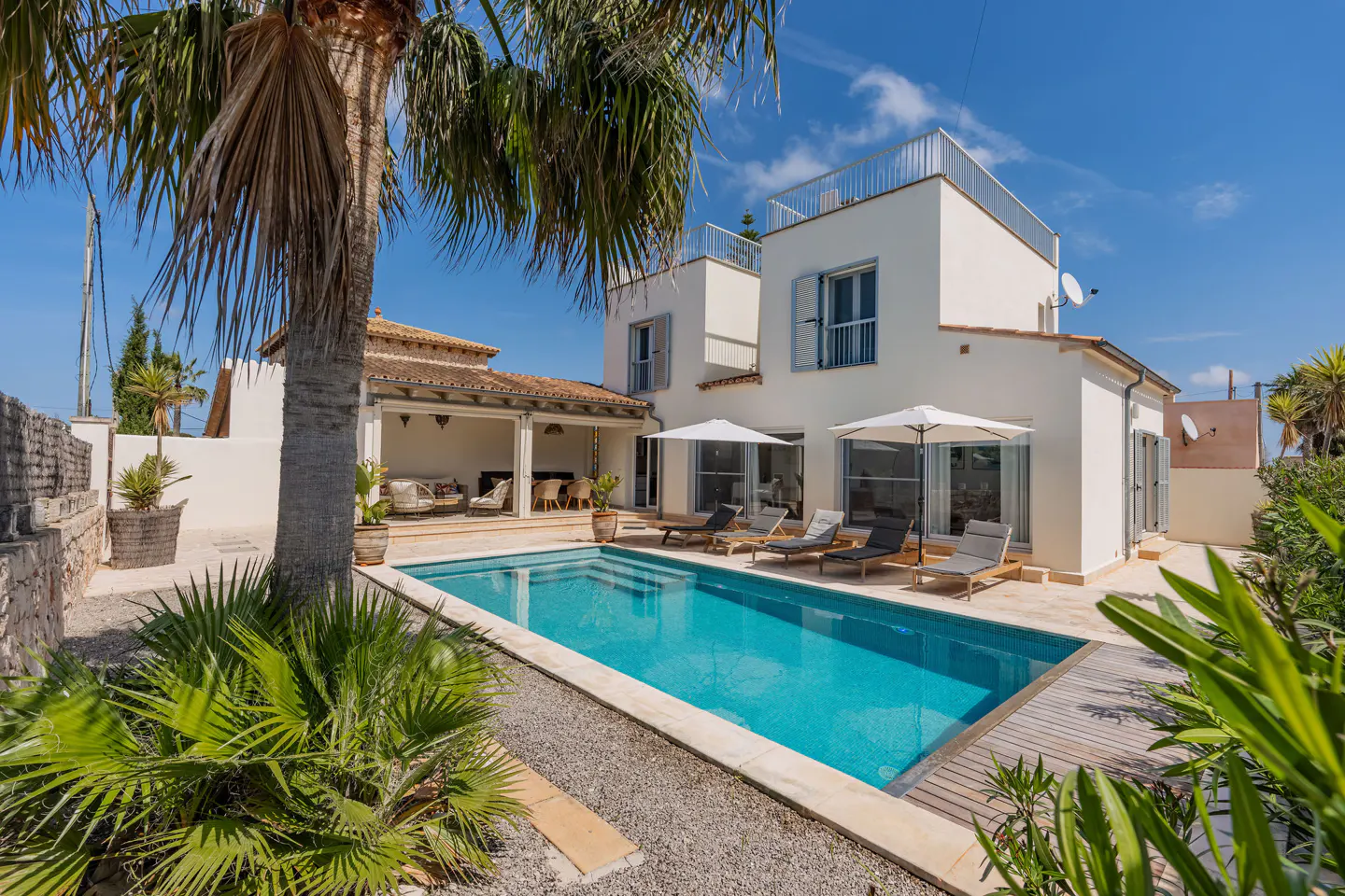 A modern white villa with a blue swimming pool, lounge chairs, and palm trees under a sunny blue sky.
