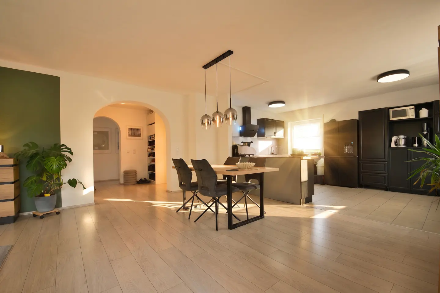 Open-concept living space with light wood floors, dining table, and black kitchen cabinets. Sunlight streams through the window.