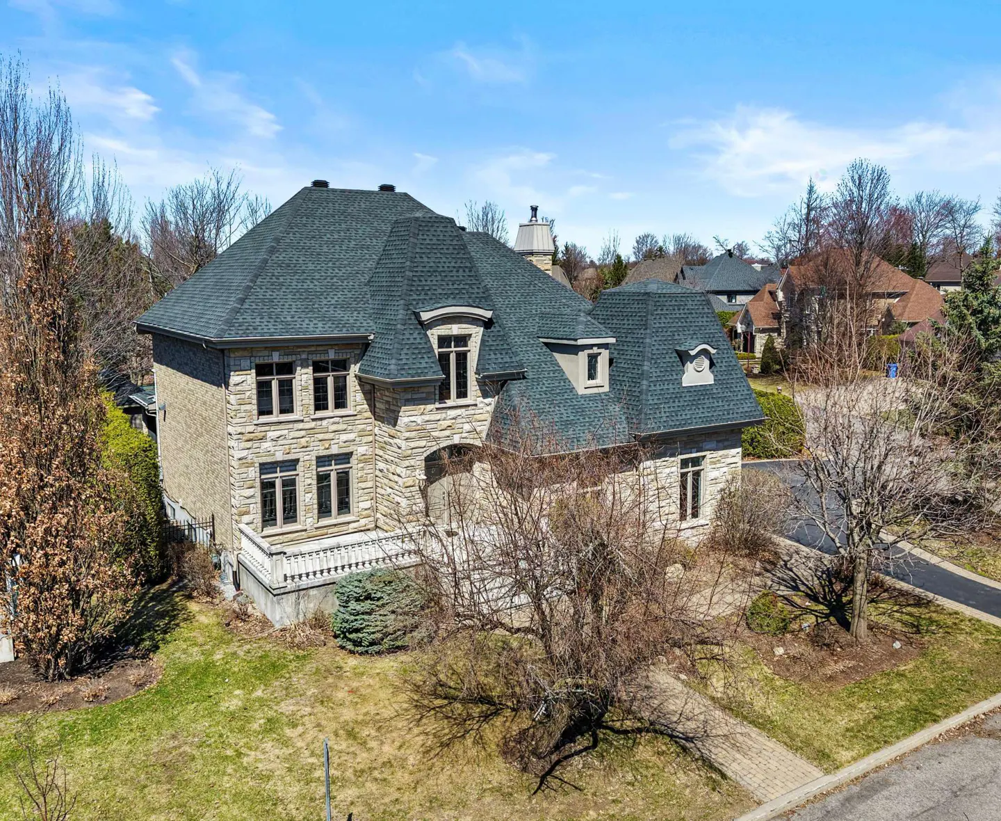 Aerial view of a large, two-story stone house with a dark gray roof and manicured lawn on a sunny day.