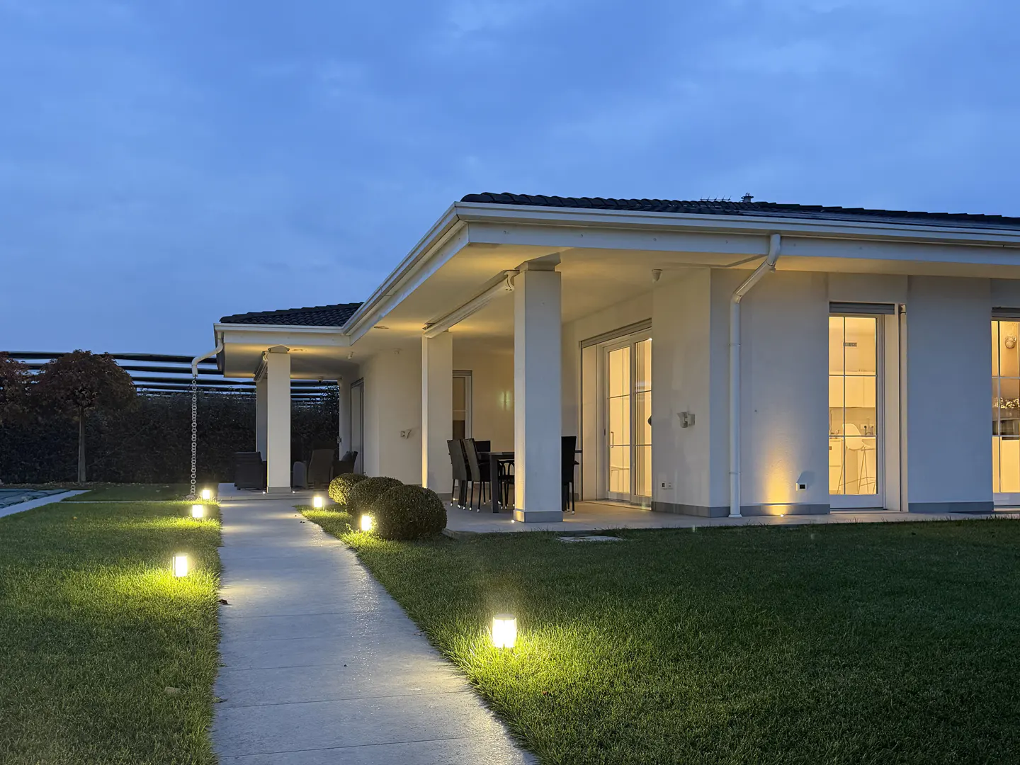 Exterior view of a modern white house at dusk, with a lit walkway, green lawn, and patio furniture.