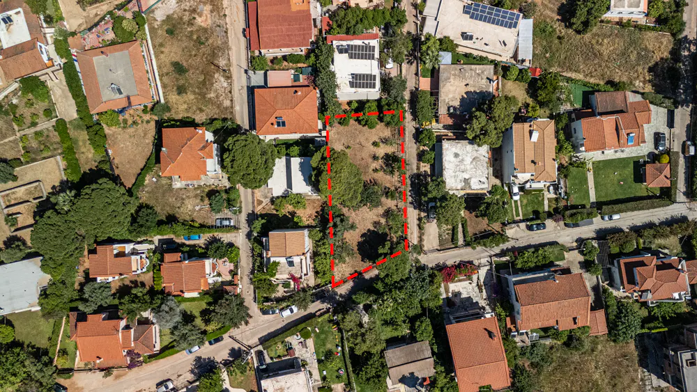 Aerial view of a vacant rectangular plot of land outlined in red, surrounded by residential houses with terracotta roofs.