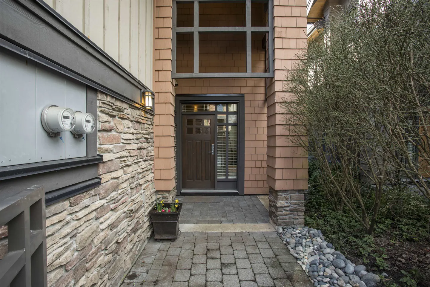 Exterior view of a home's entrance with a brown door, stone walkway, and cedar shake siding. A potted plant sits near the door.
