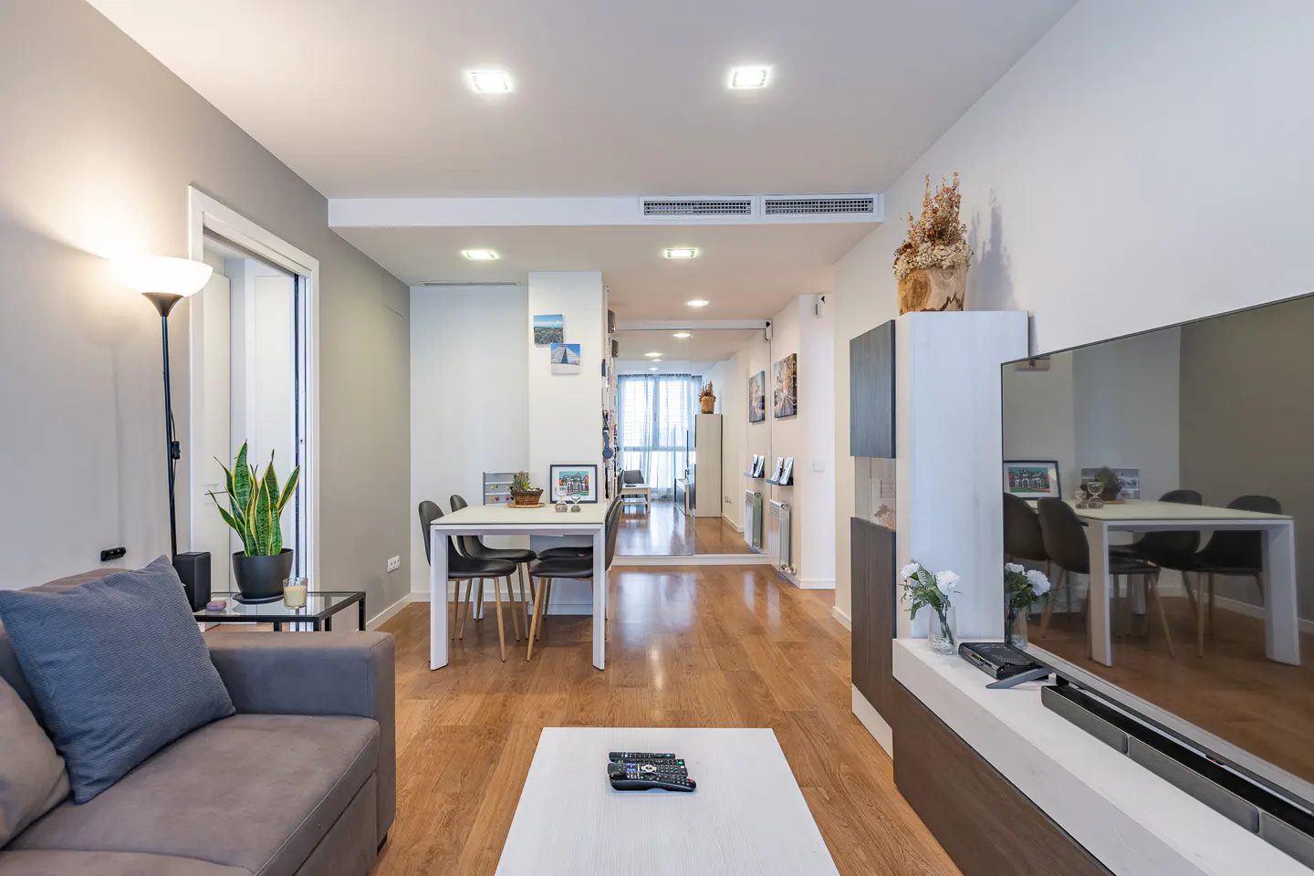 Bright, modern living room with gray sofa, white coffee table, and wood floors. Dining table and chairs visible in the background.
