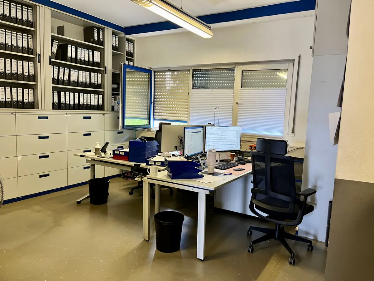 Office space with white desks, black chairs, and dual monitors. Filing cabinets line the wall, filled with black binders. Windows with blinds provide natural light.