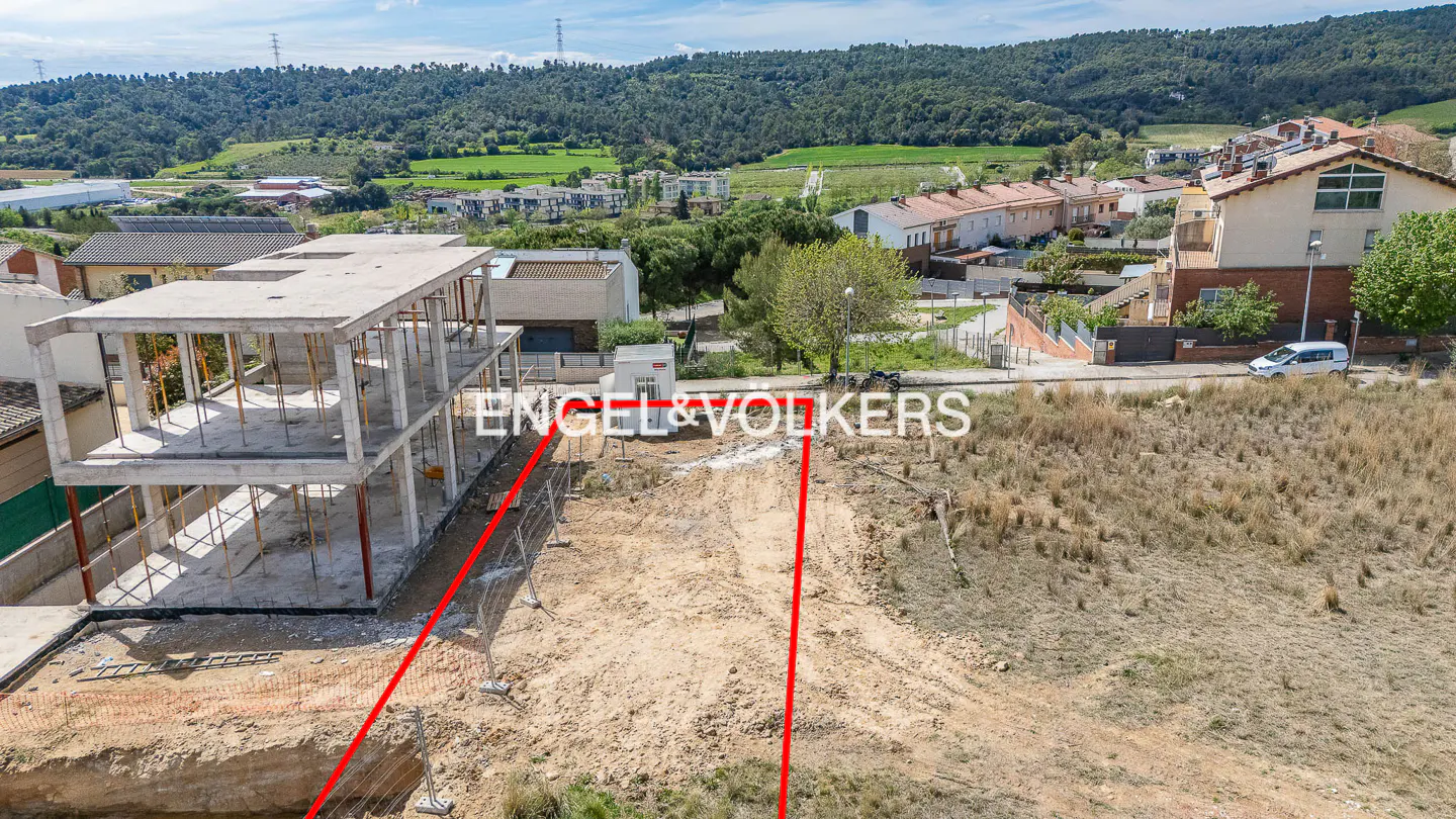 Construction site with a concrete frame building, dirt lot, and Engel & Volkers logo. Houses and green hills in the background.