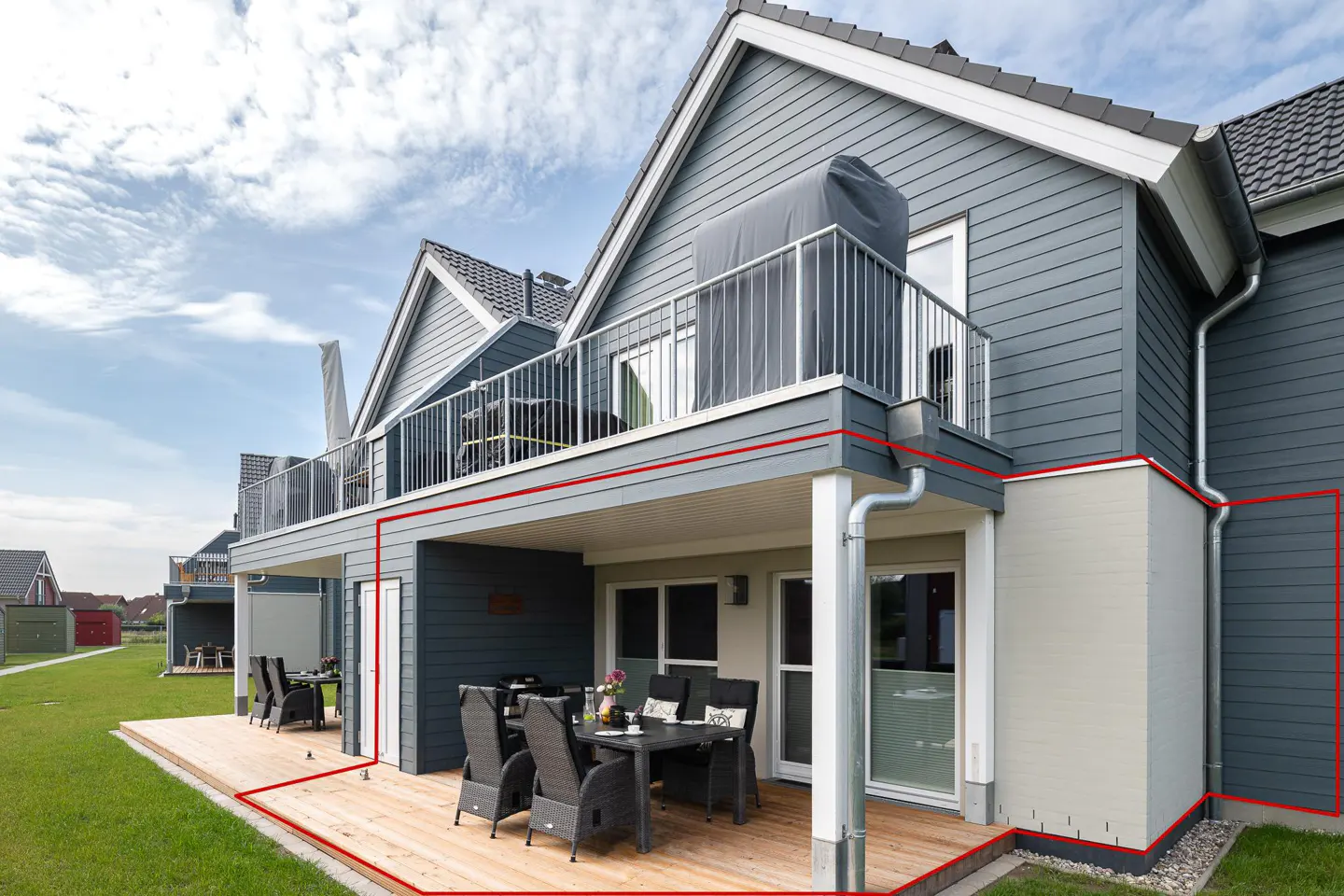 Exterior view of a two-story gray house with a wooden deck and outdoor dining set. A balcony with metal railings is on the second floor.
