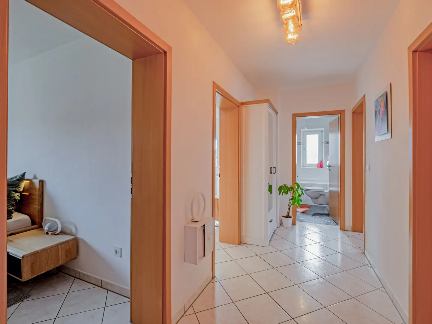Hallway with white tile floor, light walls, and wood trim. Doors lead to a bedroom and bathroom. A plant sits near the bathroom door.