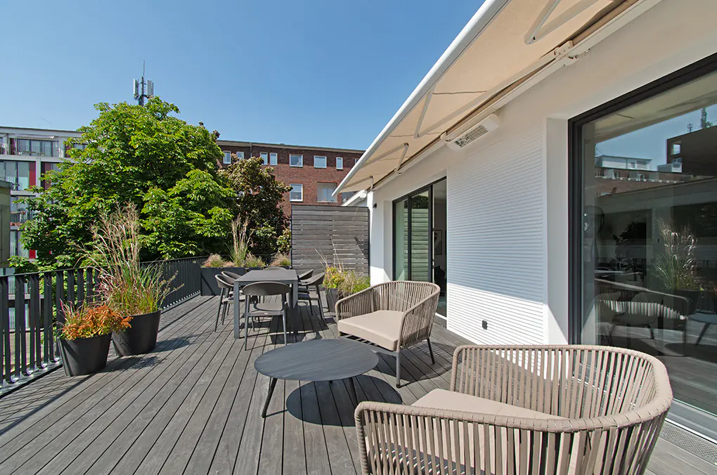 Outdoor patio with gray wood deck, table, chairs, and potted plants. A white building with a retractable awning is visible in the background.
