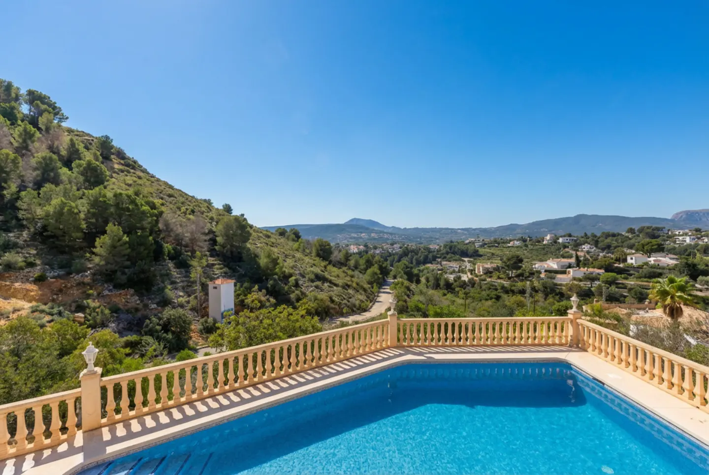 A blue tiled pool with a tan balustrade overlooks a green hillside under a clear blue sky.
