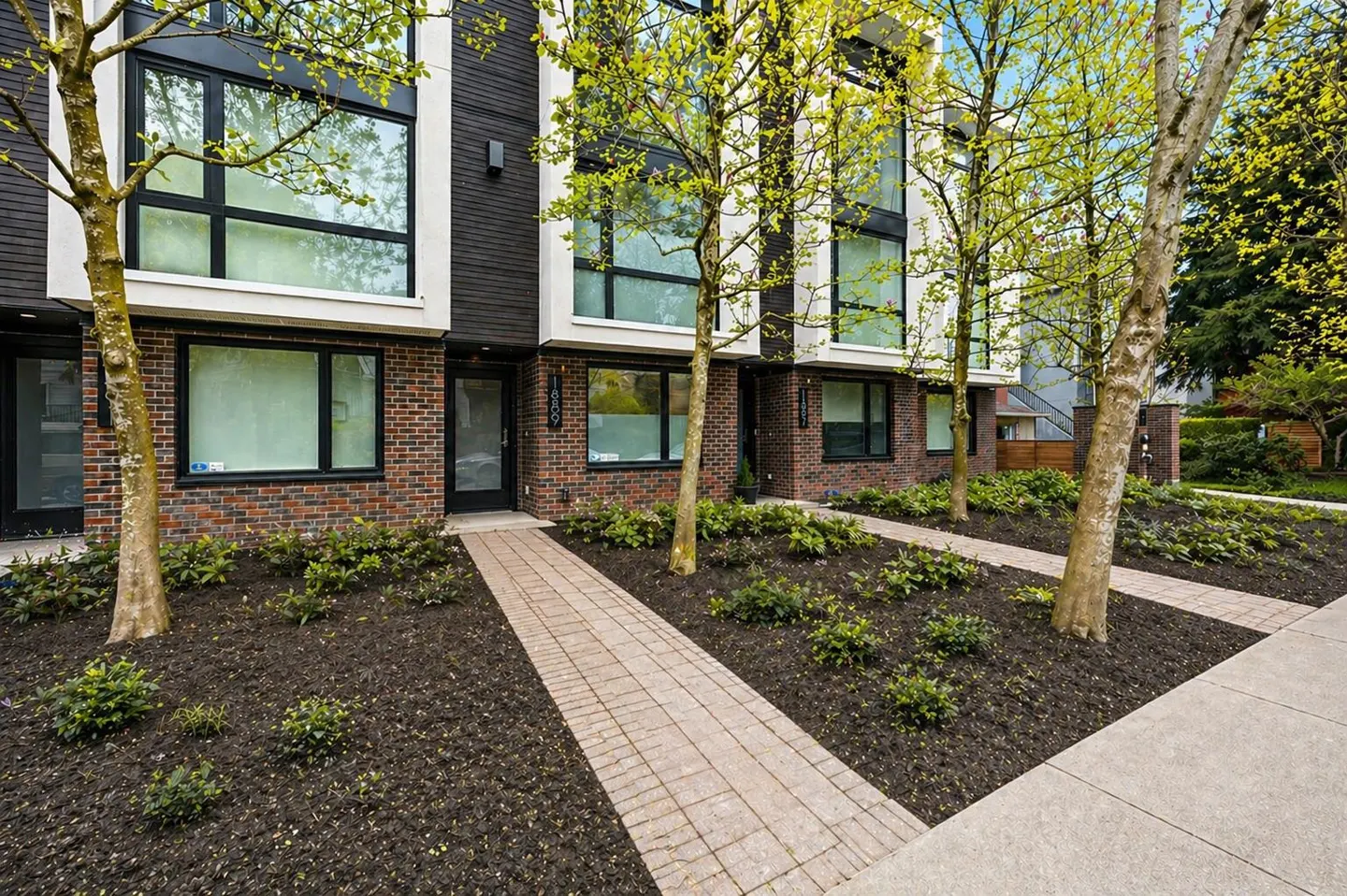 Modern townhouses with brick and dark siding, large windows, and brick walkways, surrounded by trees and landscaping.