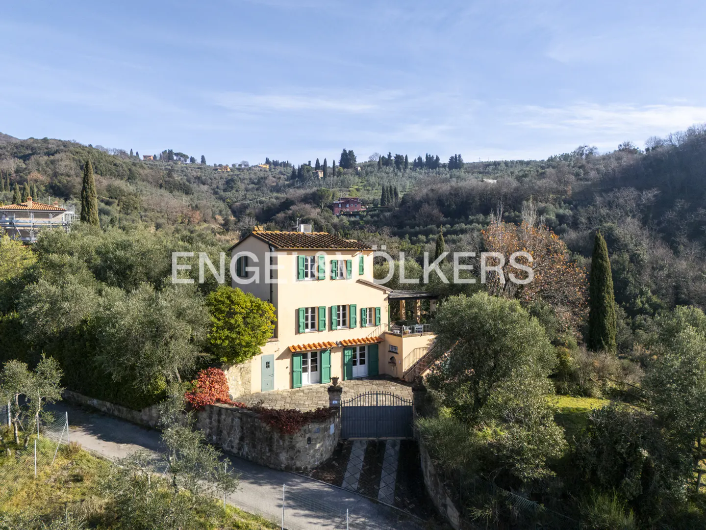 Two-story beige house with green shutters, surrounded by trees and hills under a blue sky. Stone wall and gate in front.