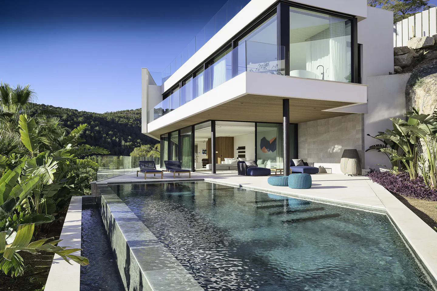 Modern white house with a pool. Glass railings and large windows offer views of the green landscape. Outdoor furniture adds a touch of luxury.