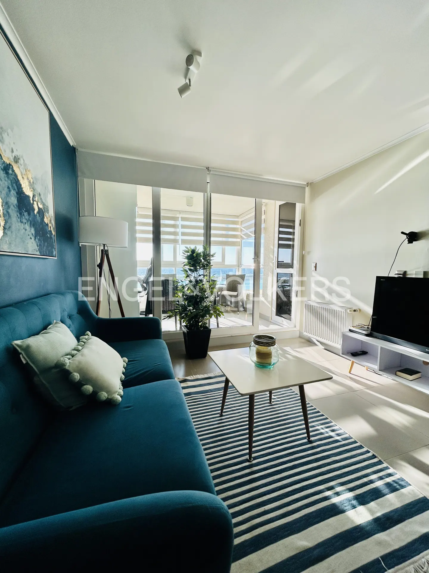 Modern living room with a teal sofa, striped rug, coffee table, and large windows overlooking a bright balcony.