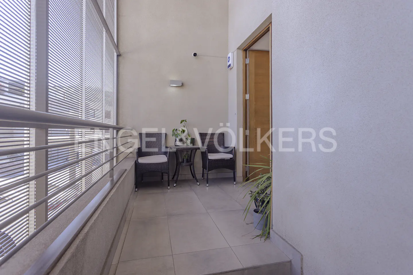 Balcony with two wicker chairs, a small table, and a plant. Blinds cover the windows. A door is visible on the right.