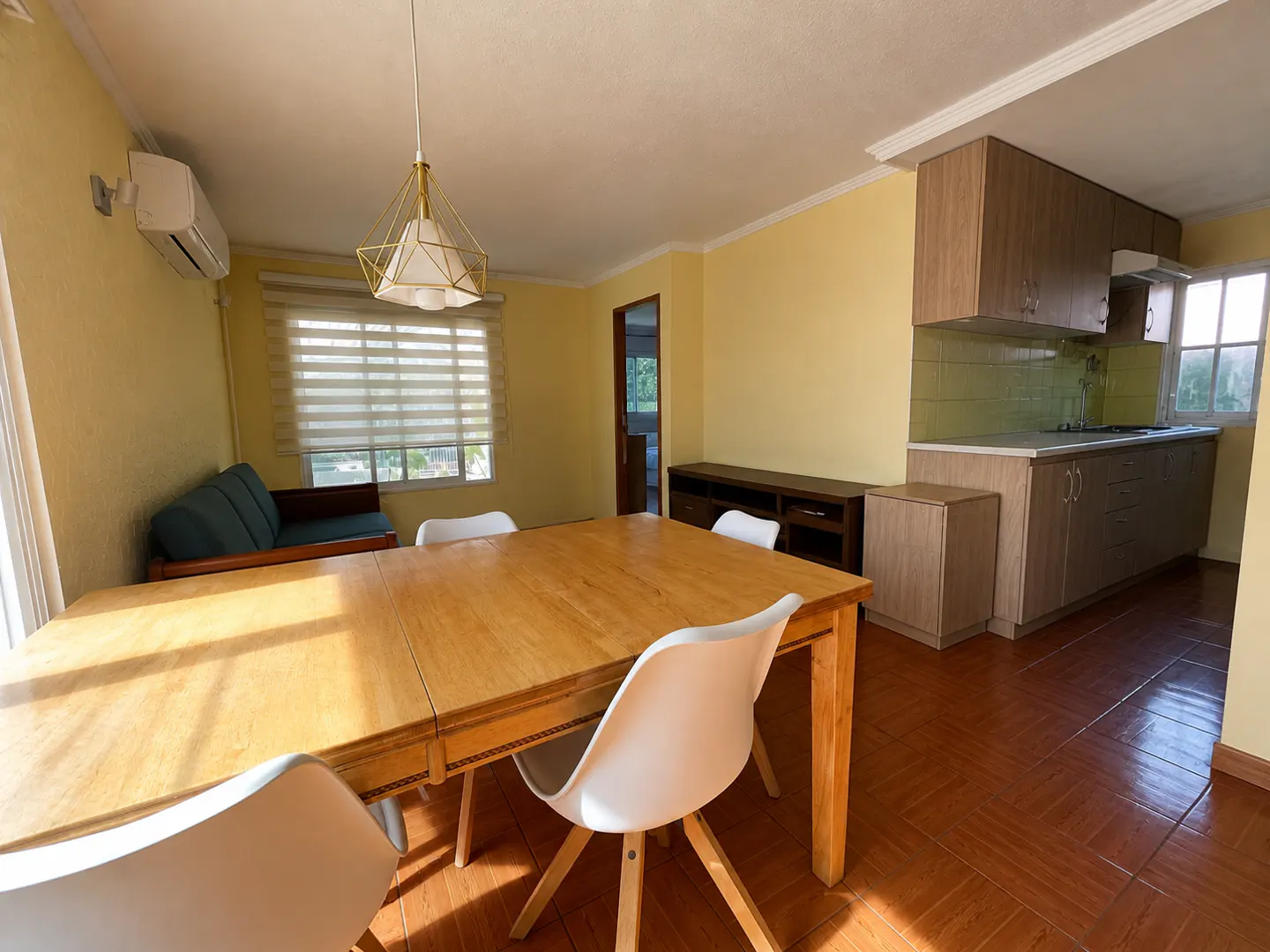 A dining room with a wooden table, white chairs, and a modern light fixture. A kitchen area is visible in the background.