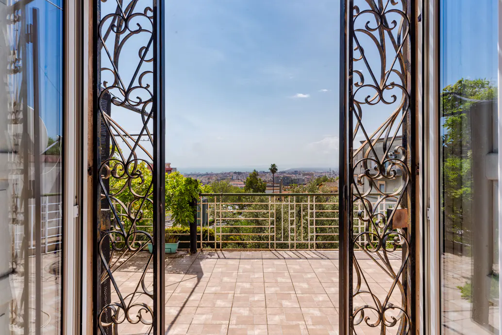 View from ornate iron doors onto a tiled balcony with a city and ocean view under a blue sky.
