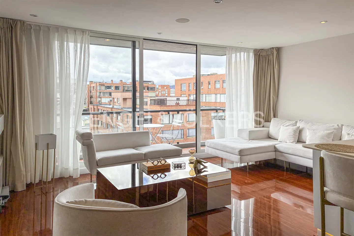 Bright living room with hardwood floors, white sofas, and a glass coffee table. Large windows offer a city view.
