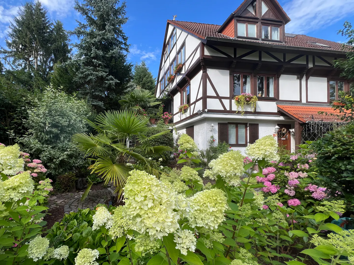 Two-story Tudor-style house with white stucco and dark brown timber framing, surrounded by lush green landscaping and hydrangeas.