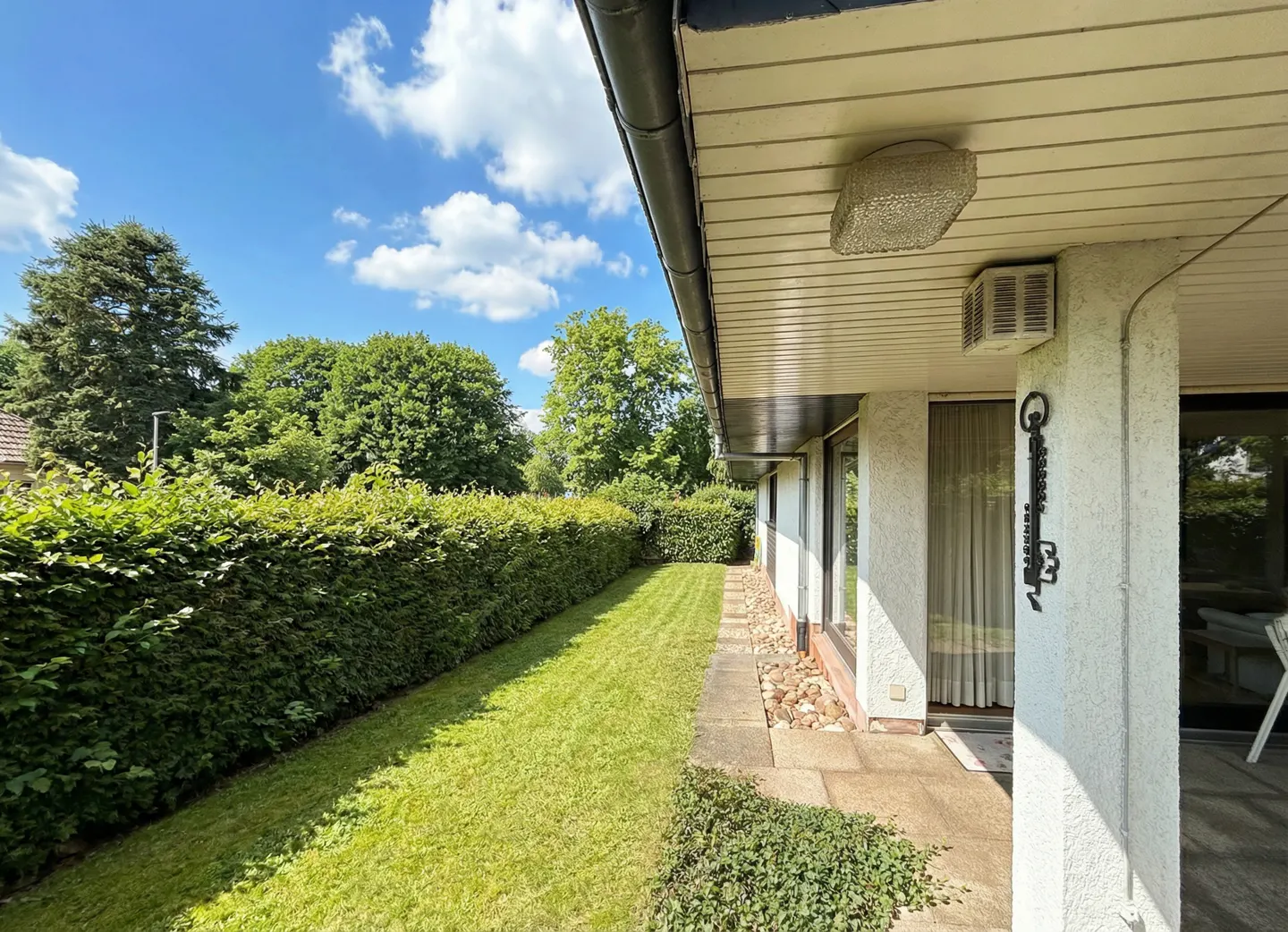 Exterior view of a house with a green lawn, hedge, and trees under a blue sky with white clouds. The house has a white exterior with a covered porch and a decorative metal key hanging on a pillar.