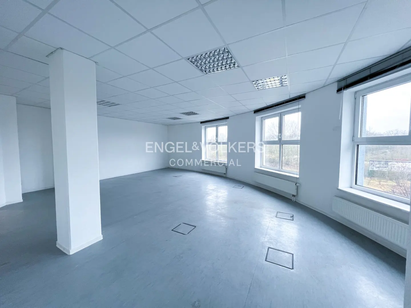 Empty, bright commercial office space with white walls, grey flooring, drop ceiling, and large windows.
