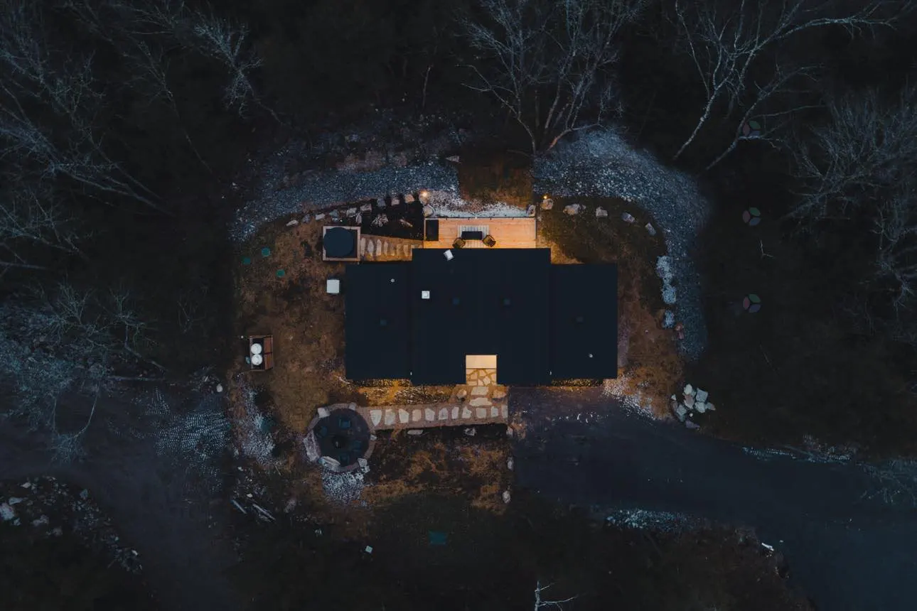 Aerial view of a dark-roofed house surrounded by trees, with a stone path leading to the entrance and a fire pit nearby.