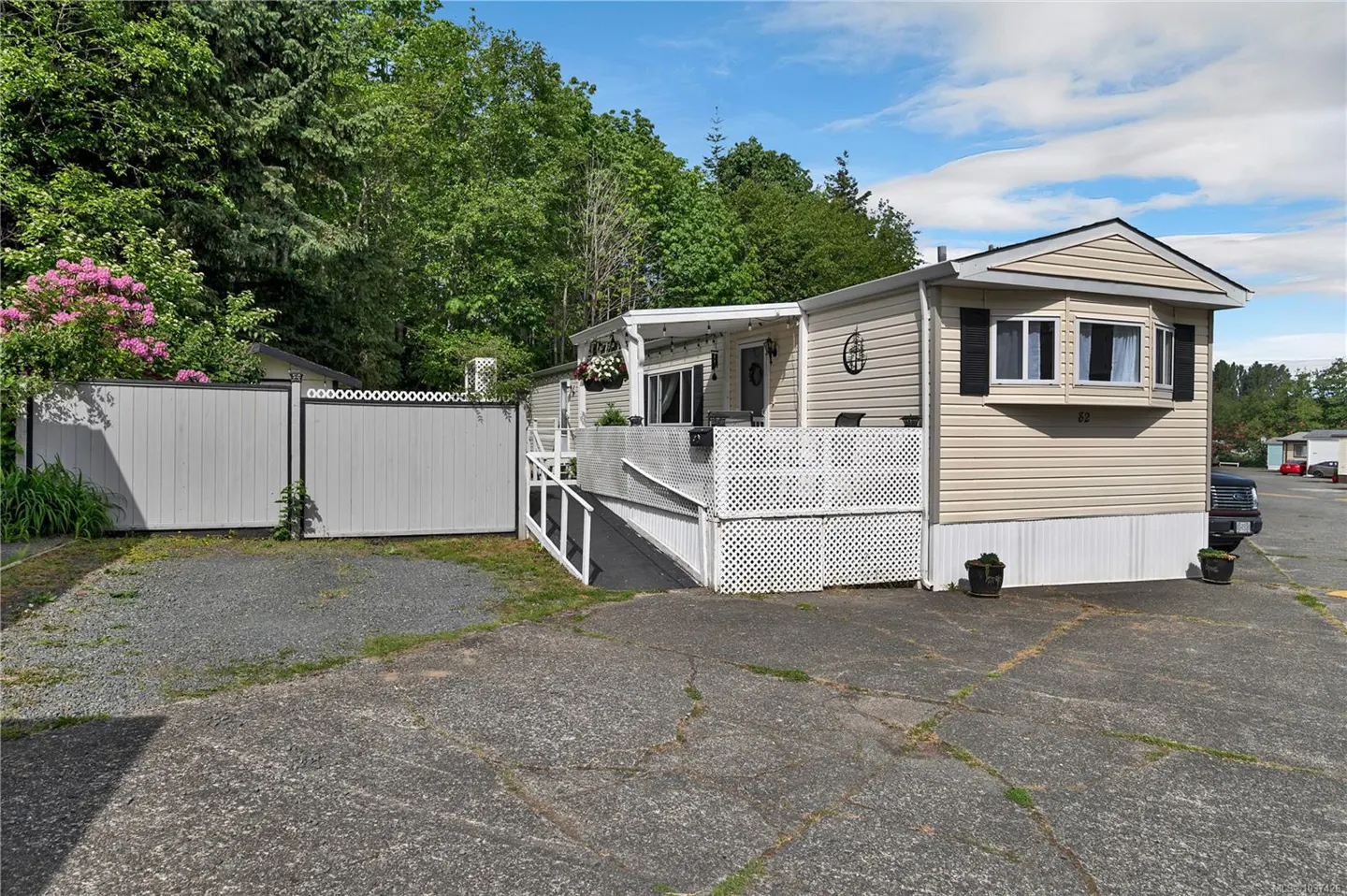 A tan mobile home with a white lattice porch and a gray fence, surrounded by trees and a cracked asphalt driveway.