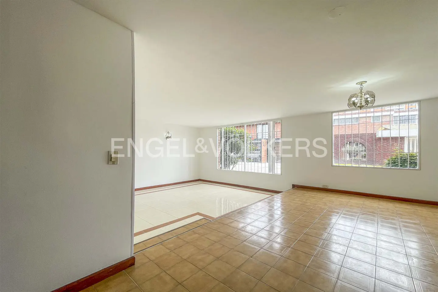 Bright, empty living room with tile floors, white walls, and large windows with views of a brick building and greenery.