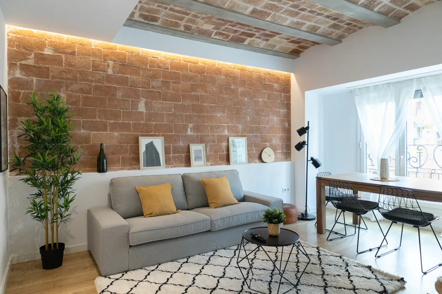 Modern living room with a grey sofa, yellow pillows, brick wall, vaulted ceiling, and a dining area with black chairs.