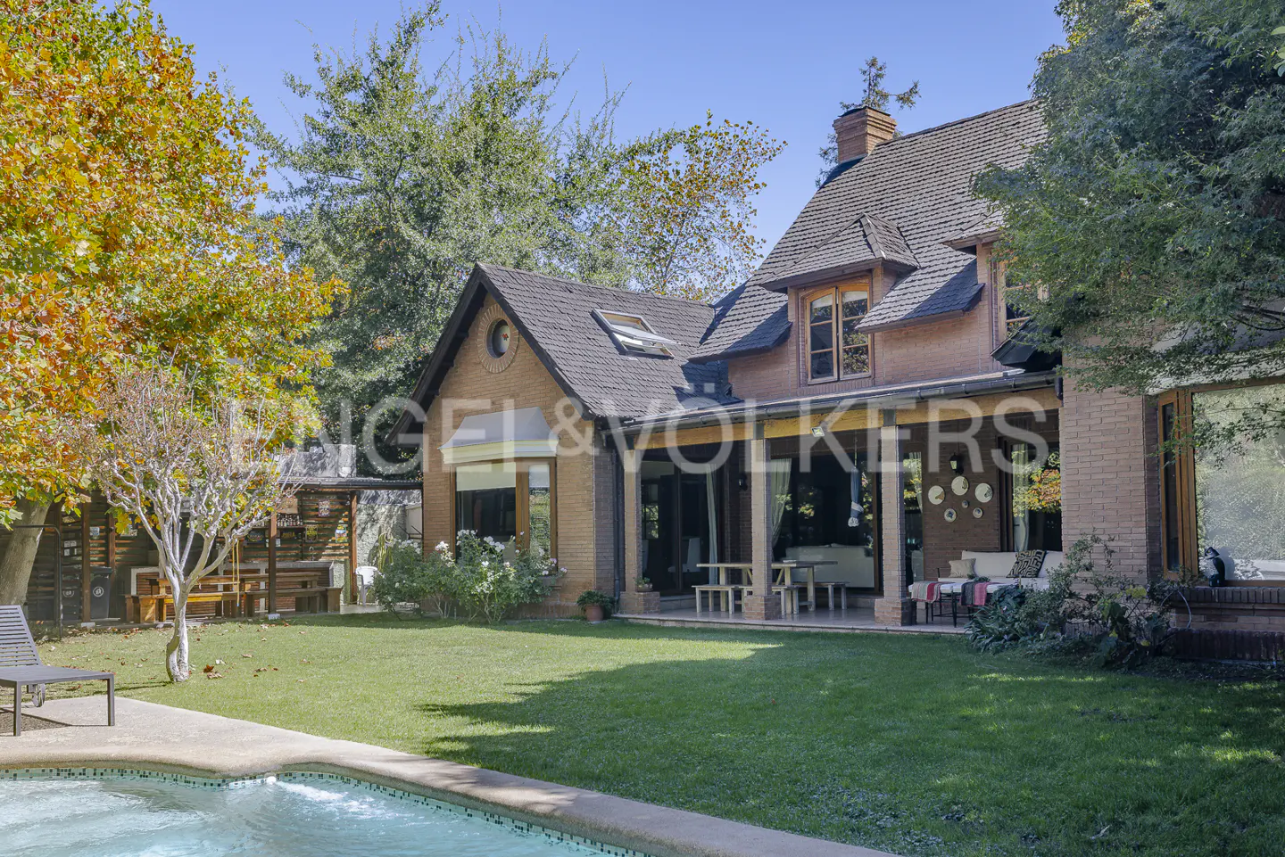 Exterior view of a brick house with a pool, lawn, and trees under a blue sky. The house has a covered patio and the Engel & Volkers logo.
