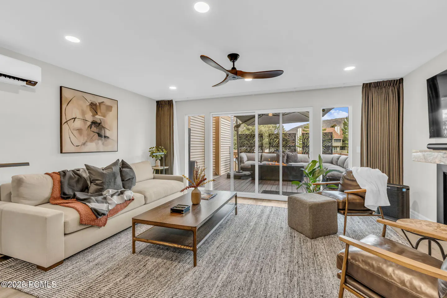 Bright living room with a beige sofa, brown leather chairs, and a wooden coffee table on a gray rug. Sliding glass doors lead to an outdoor patio.