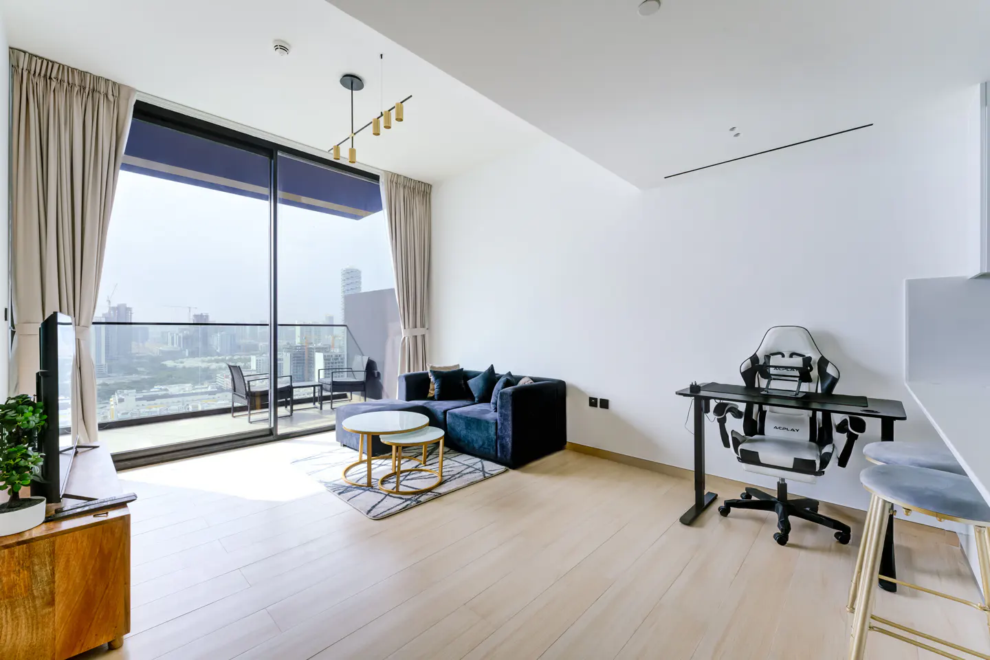 Bright apartment interior with a blue sofa, round tables, a gaming chair and a balcony overlooking a city.