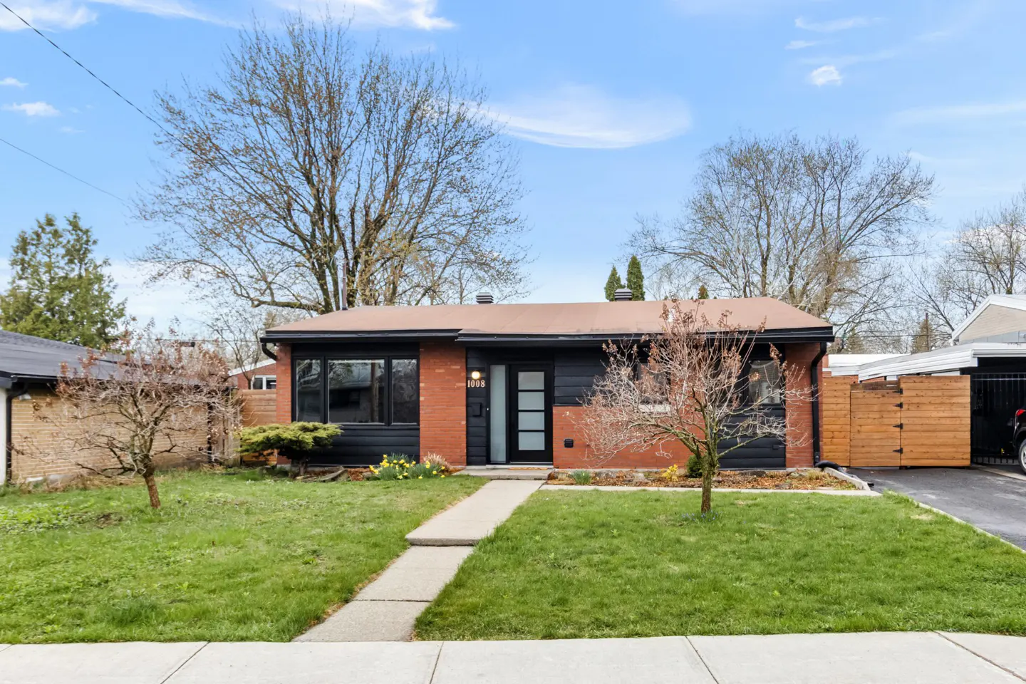A single-story house with a black and brick exterior, a brown roof, and a green lawn under a blue sky.