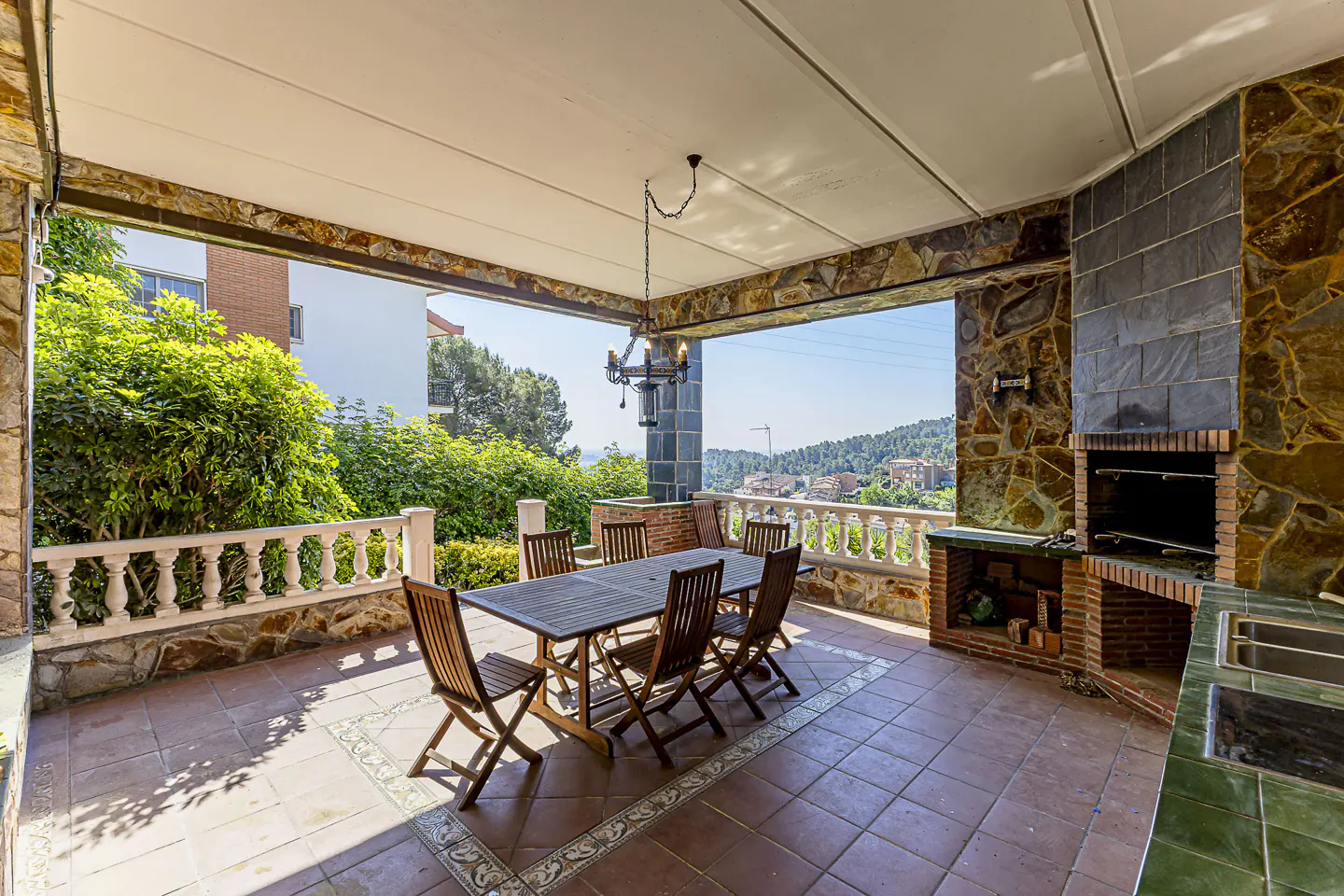 Covered patio with stone walls, tile floor, and a dining table with six chairs. A built-in brick grill and sink are on the right. Balcony overlooks a green landscape.