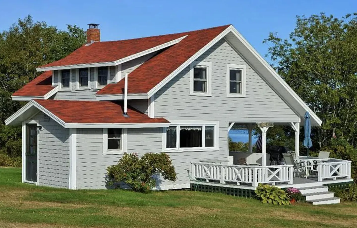 A two-story gray house with a red roof and white trim sits on a green lawn under a blue sky. A white porch is visible on the right.