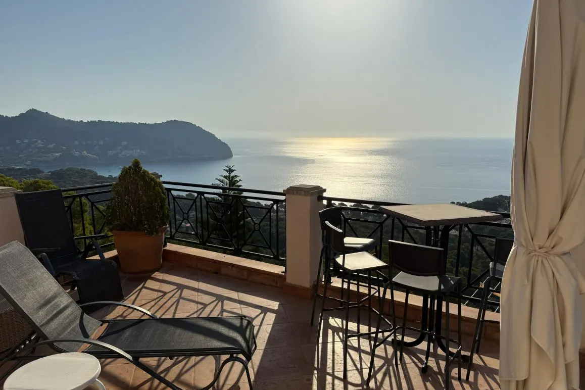 Balcony with lounge chairs, table, and chairs overlooking the ocean and a distant mountain range on a sunny day.