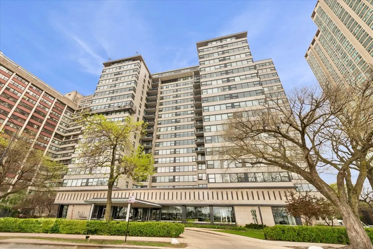 Exterior view of a tall, gray apartment building with many windows and a covered entrance. Trees are in the foreground.