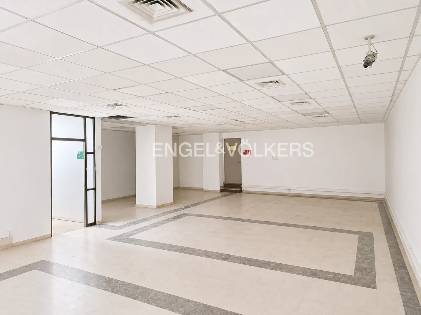 Empty office space with white walls, tiled floor, and drop ceiling with a security camera.