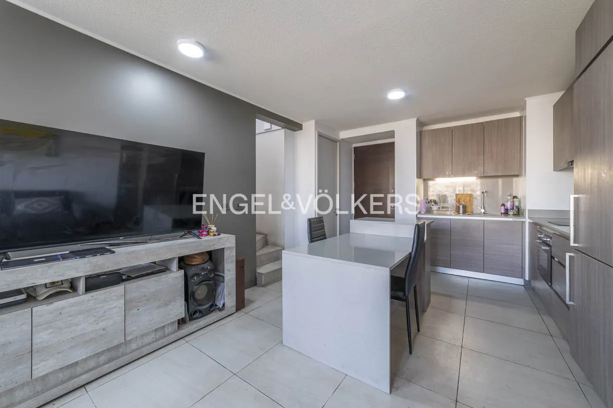 A modern living space with a gray wall, TV, and a white kitchen island with black chairs. Gray cabinets and appliances are visible in the kitchen area.
