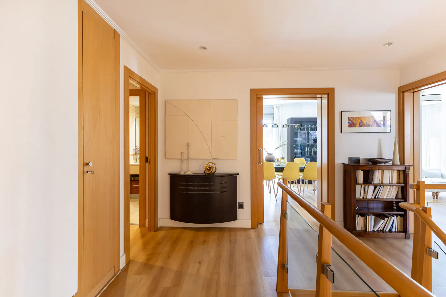 Hallway with light wood floors, white walls, and wood trim. A black radiator cover sits below a white abstract painting. Dining room with yellow chairs visible through doorway.