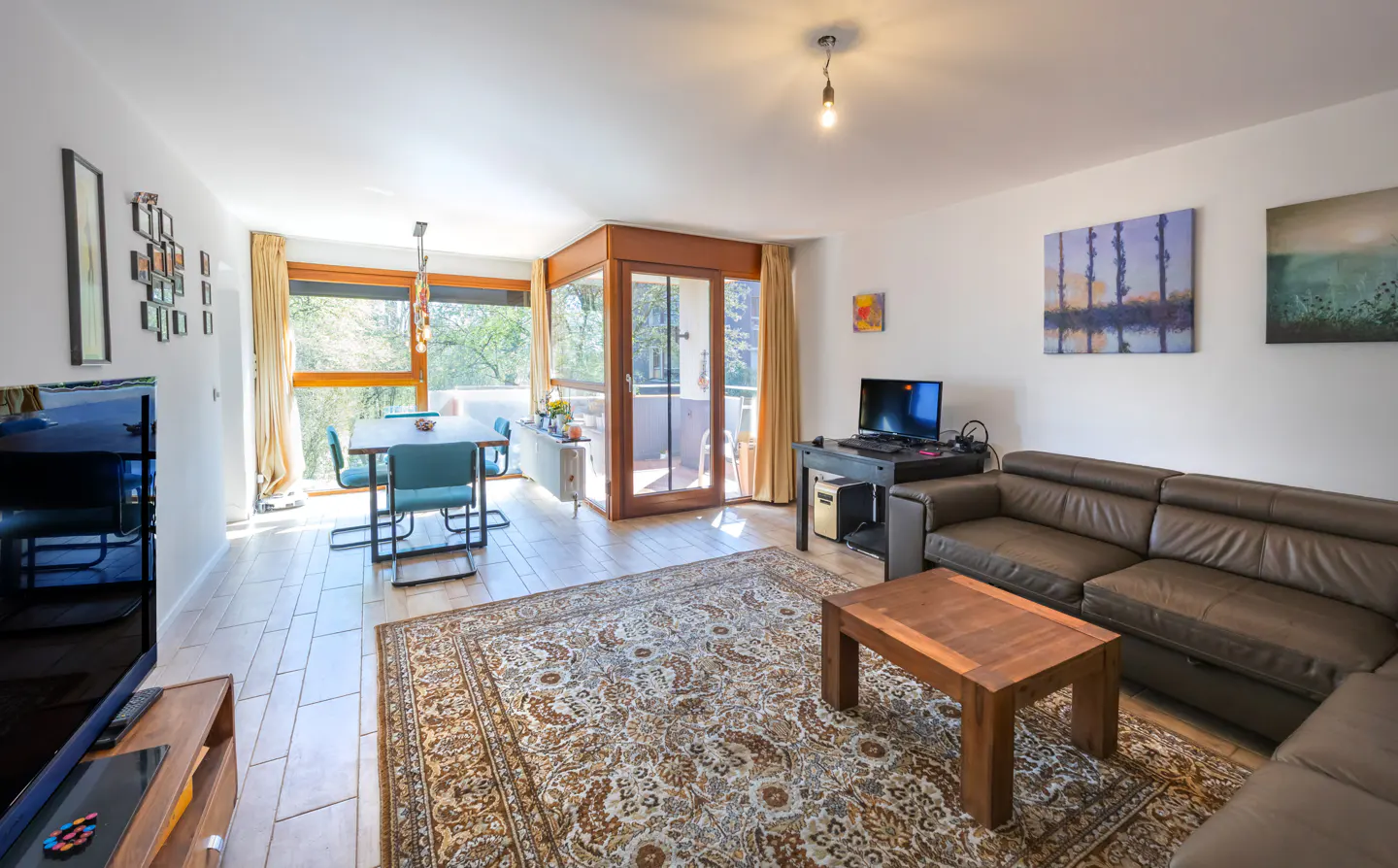 Bright living room with a brown leather sofa, wooden coffee table, and patterned rug. A dining table and balcony are visible in the background.