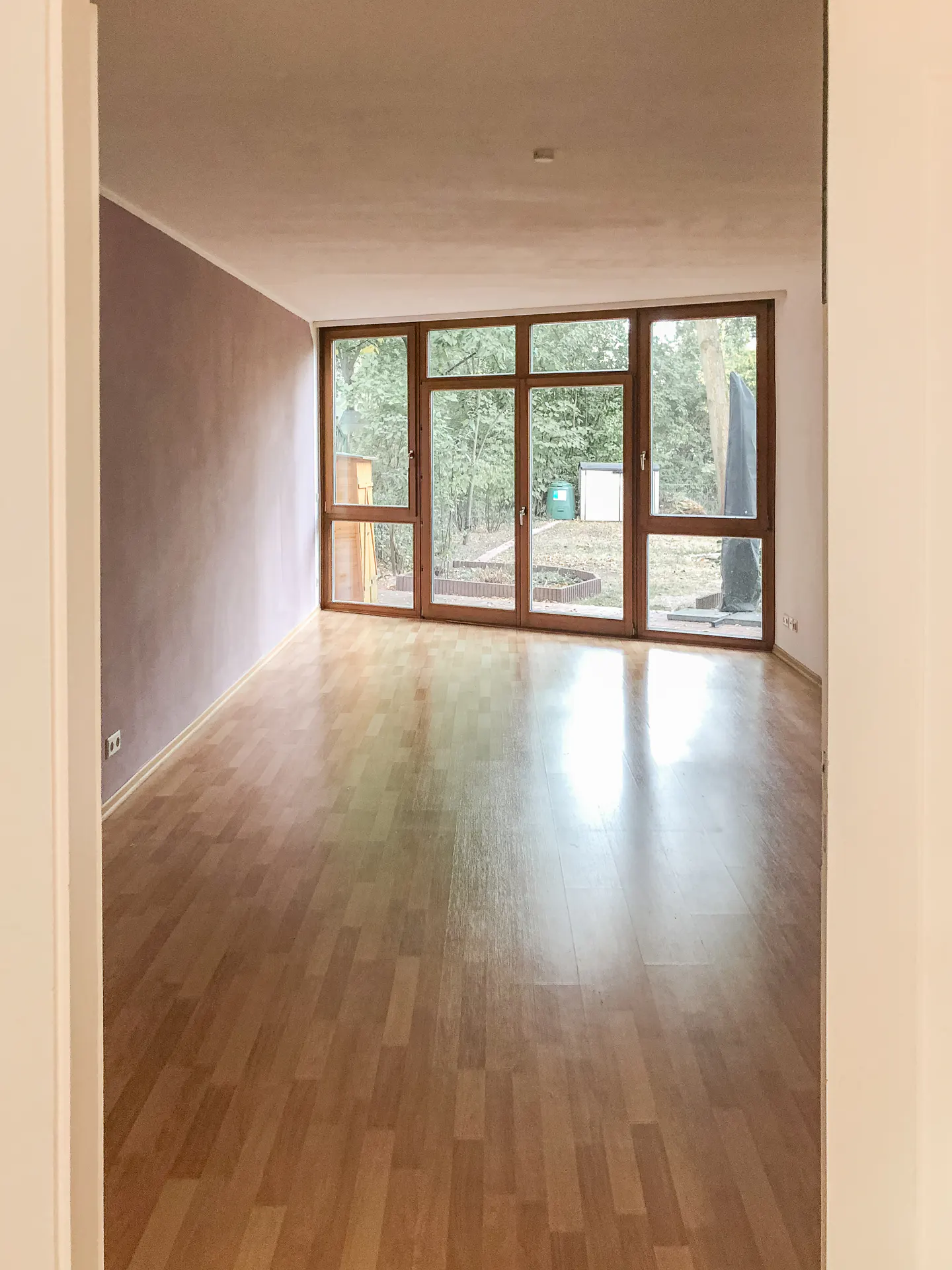Empty room with light wood floors, a lilac wall, and large windows overlooking a green yard.