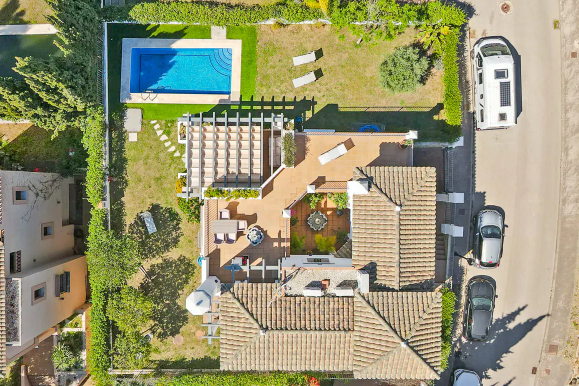 Aerial view of a property with a pool, patio, and garden, surrounded by lush greenery and parked cars.