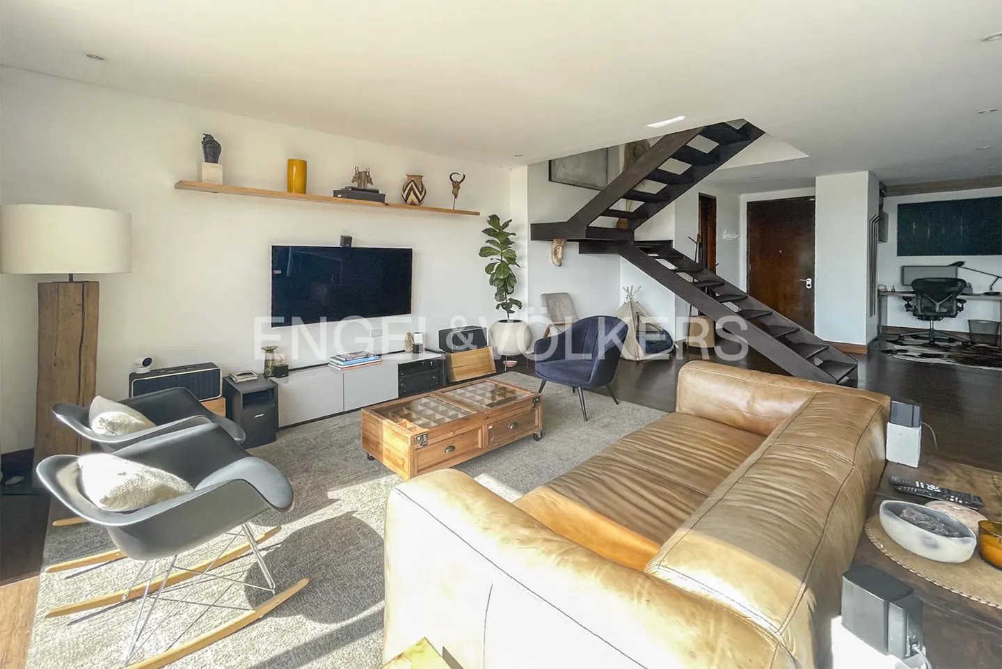 Bright living room with a brown leather sofa, rocking chairs, a wooden coffee table, and a black staircase leading to the second floor.