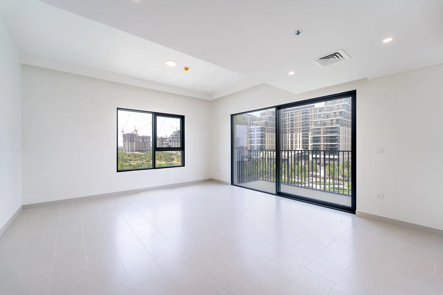 Bright, empty room with white walls and tile floor. Black-framed window and sliding glass doors offer city views.