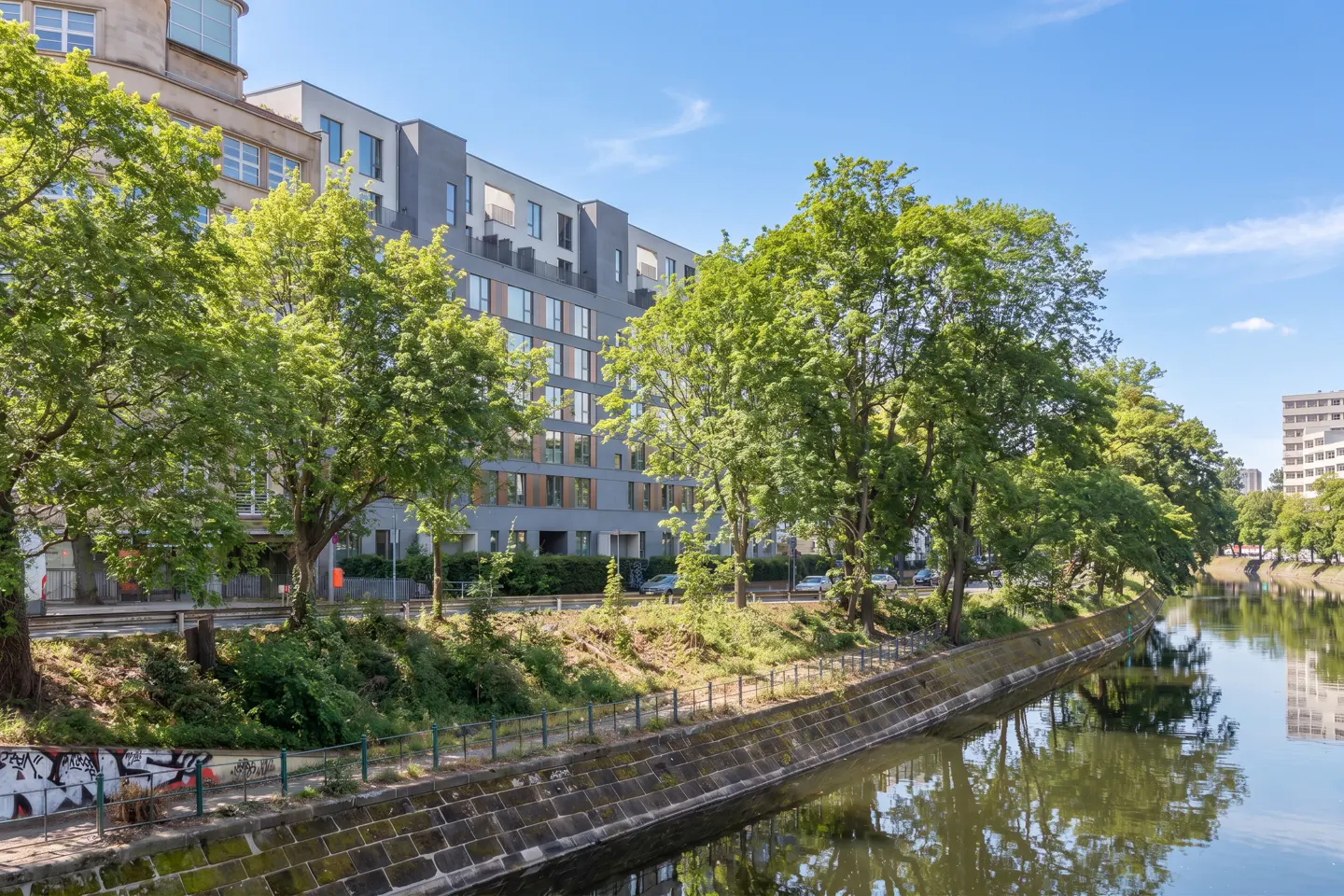 A modern gray apartment building sits beside a canal lined with green trees under a blue sky.