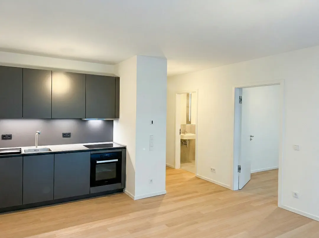 Bright, modern apartment with gray kitchen cabinets, stainless steel sink, and light wood floors. Open doorway leads to a bathroom.