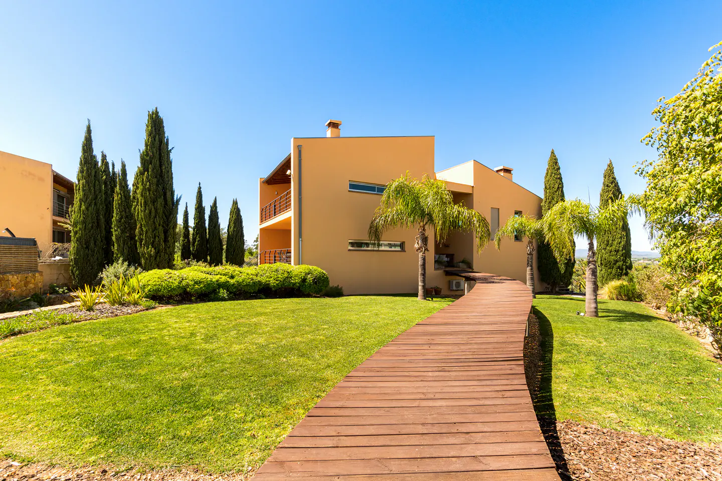 Exterior view of a tan two-story house with a wooden walkway, green lawn, palm trees, and tall cypress trees under a clear blue sky.