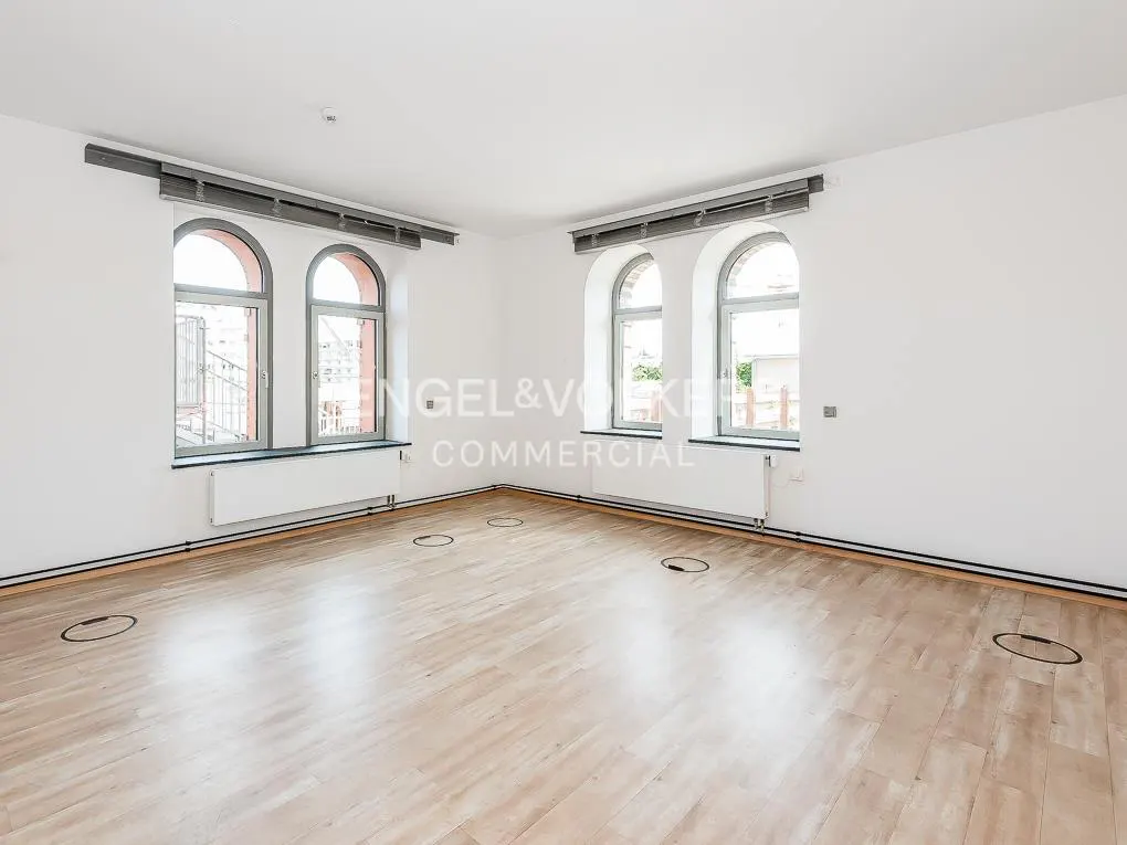 Bright, empty room with light wood floors, white walls, and arched windows. Radiators are under the windows. Floor outlets are visible.