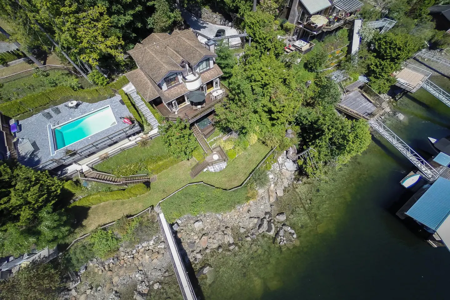Aerial view of a luxury waterfront home with a pool, dock, and lush green landscaping. The house has a brown roof and multiple decks.