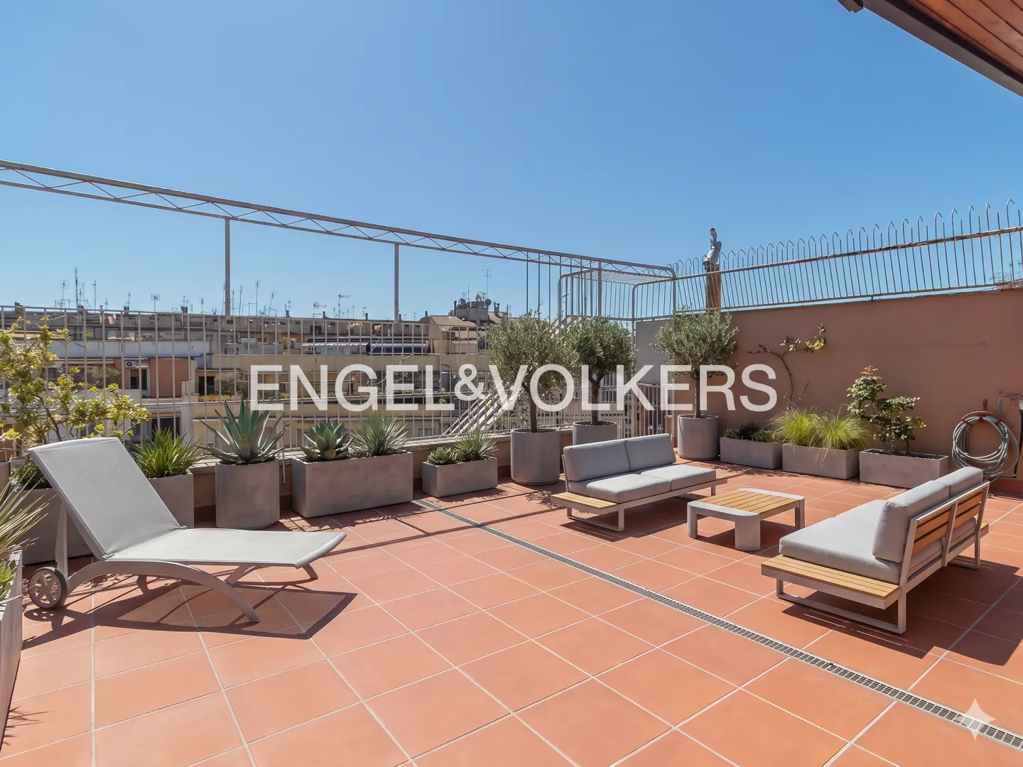 Rooftop terrace with terracotta tiles, lounge chairs, sofas, and potted plants under a clear blue sky. Metal railing surrounds the space.